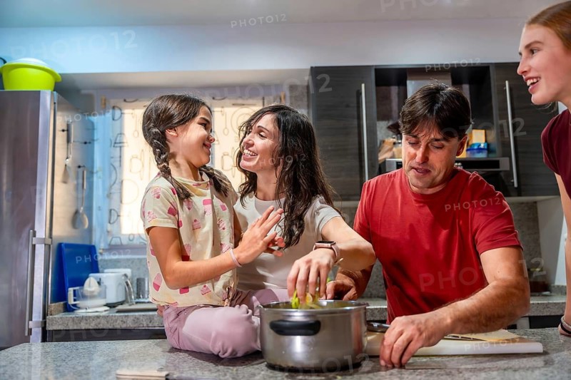 Family including parents and children standing and sitting in a bright kitchen, happily engaging in meal preparation, laughing and cooking together in a home environment