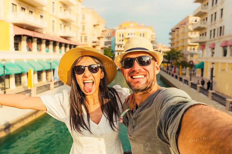 Happy tourist couple taking selfie Qanat Quartier Doha. Woman sticking tongue out, man smiling, straw hats, sunglasses. Venetian style canals, colorful buildings background
