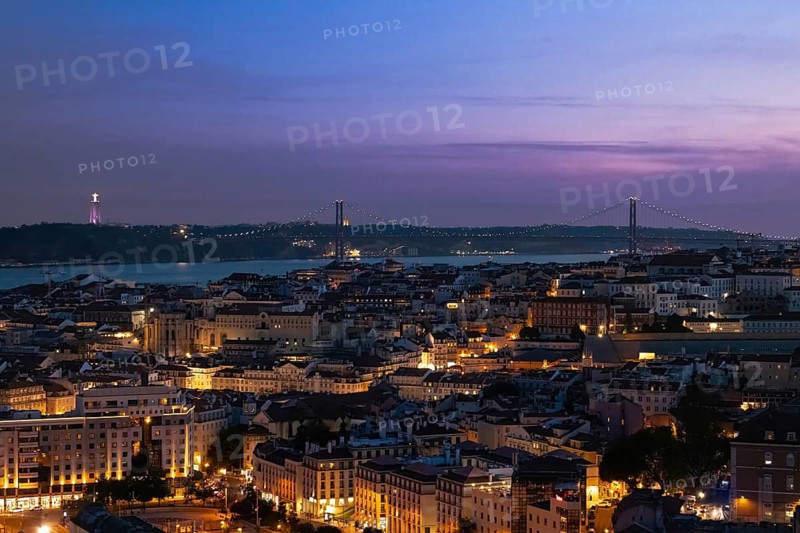 High angle panoramic view of Lisbon city skyline at dusk with illuminated streets, historic architecture the Tagus river and the suspension bridge