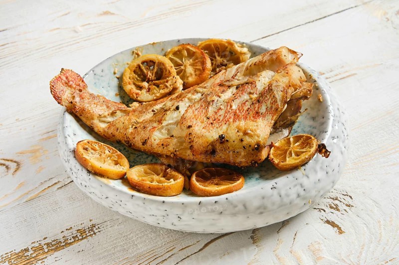 Roasted whole red snapper is placed on plate along with roasted lemon slices. Fish ready for serving on simple wooden table. Meal looks fresh and appetizing