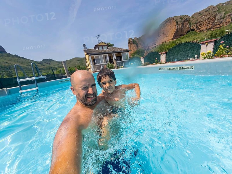 Father holding his son while playing and splashing in a bright blue swimming pool on a private property with mountains and a house in the background on a sunny day
