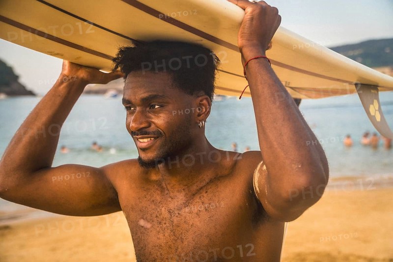African man carrying a surfboard on his head walking along a sandy beach, smiling while looking towards the ocean with swimmers in the background during a summer day