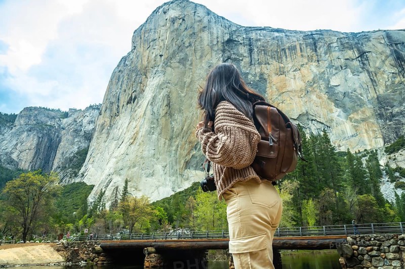 Woman traveler with a backpack and camera standing by a river, observing the massive granite cliffs of el capitan in yosemite valley, embracing the beauty of nature and adventure