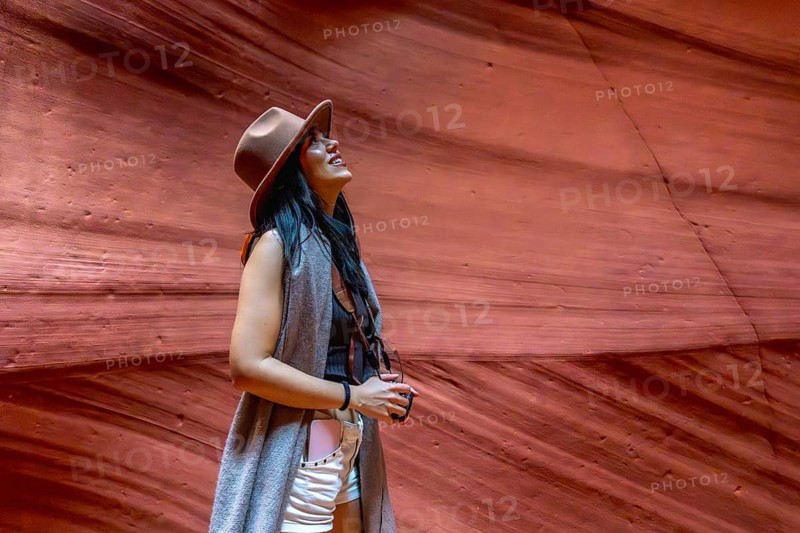 Woman standing inside antelope canyon exploring the geological beauty of the slot canyon, wearing a hat and light clothing while looking up at the light reflecting on the unique sandstone formations