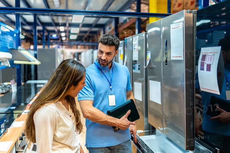 Salesman helping a young woman examine an open refrigerator in a warehouse style home improvement store, discussing features and specifications for her new kitchen appliance