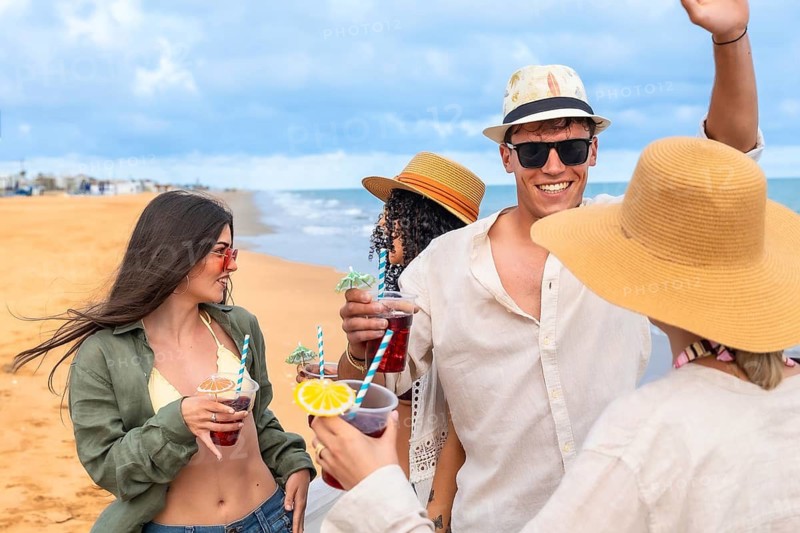 Young friends toast cocktails on a sunny sandy beach at sunset, smiling and celebrating summer vacation together, relaxed and carefree by the ocean waves