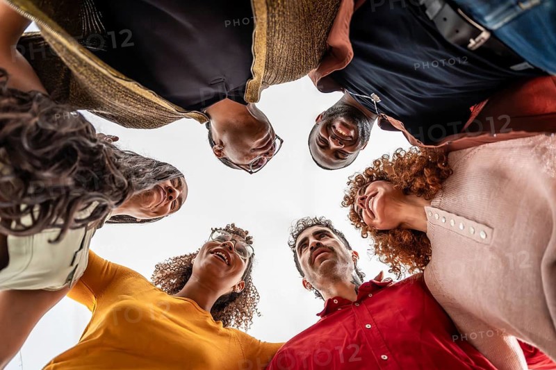 Diverse group of smiling people huddling together in a circle, looking down with arms around each other, fostering a sense of community, unity, and support from a low angle perspective