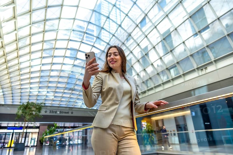 Young businesswoman standing in a modern transportation hub, smiling while looking at her smartphone, connecting with others or capturing a moment in a busy urban environment