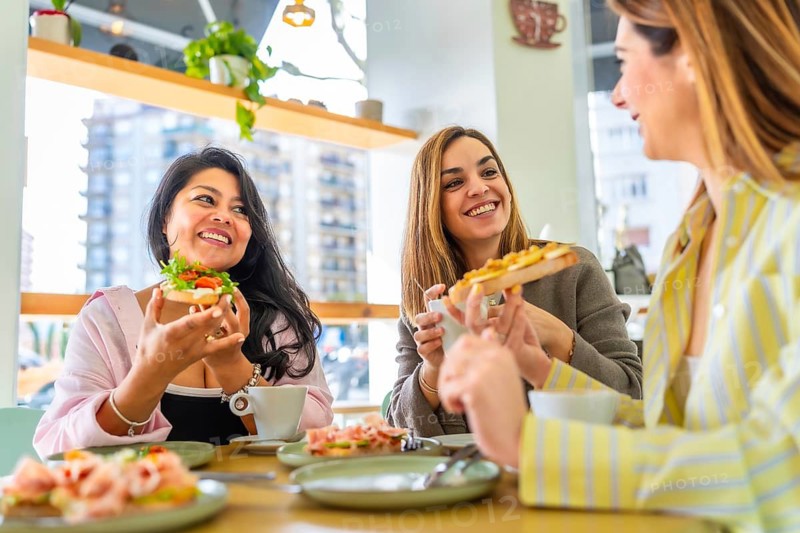 Diverse group of female friends smiling and conversing while eating delicious avocado toast and open sandwiches during a joyful friendly gathering at a modern restaurant