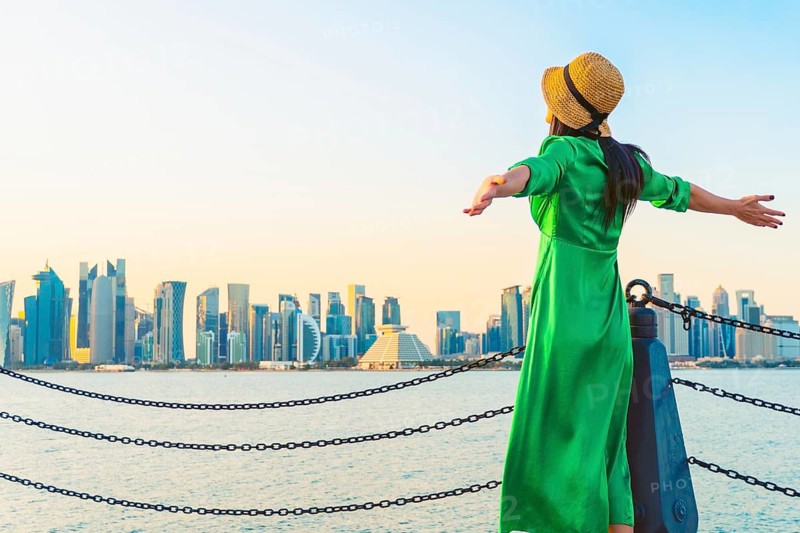 Woman traveler visitor standing with open arms at waterfront facing Doha skyline across bay symbolizing freedom urban lifestyle travel Middle East modern city concept
