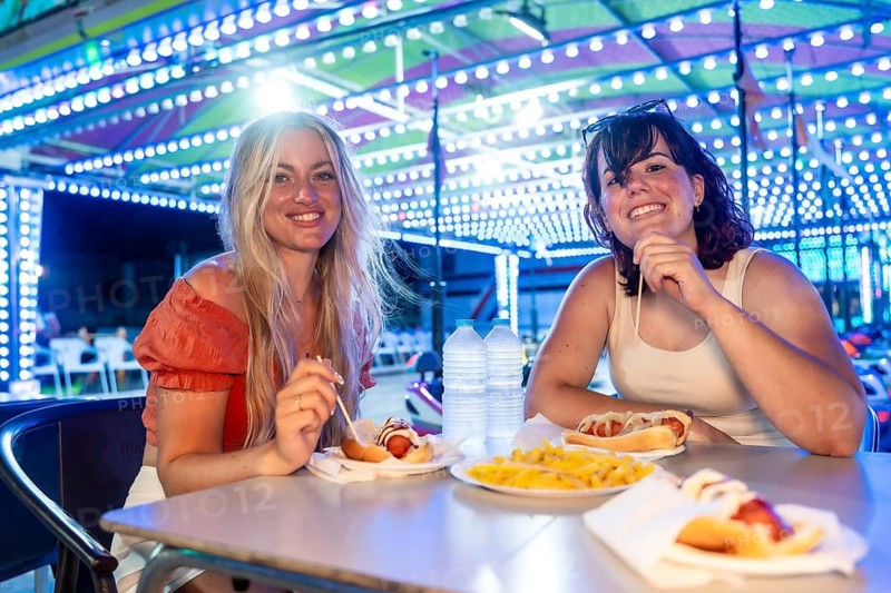 Two smiling young adult women sitting at a table, eating hot dogs and french fries, enjoying time together at a vibrant funfair with bright lights after sunset