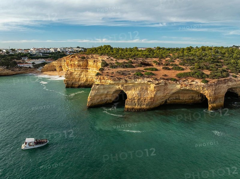 Drone view of Benagil Cave cliffs in Algarve, Portugal, with a tourist tour boat, limestone sea caves, turquoise Atlantic water, and waves