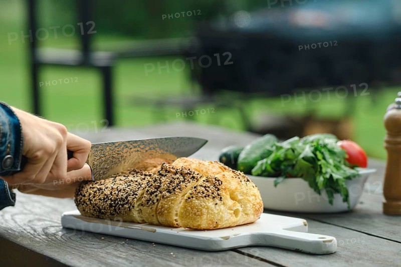 Person uses knife to slice fresh bread on wooden table outside. Grill is visible in background. Vegetables are on the table. Weather appears clear and suitable for outdoor cooking
