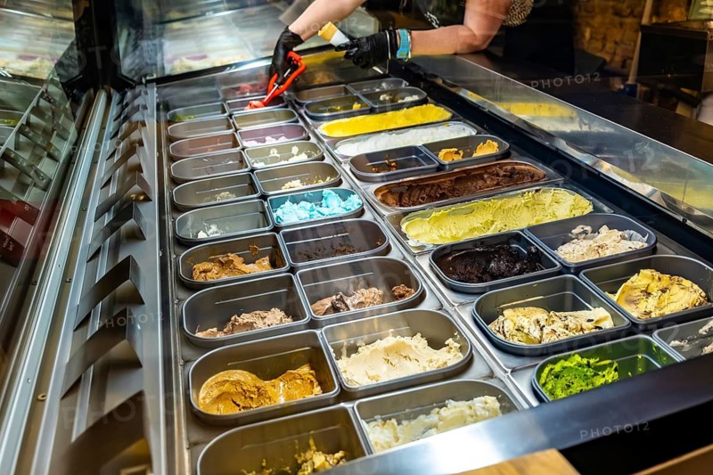 Person wearing gloves scooping vibrant gelato flavors from a refrigerated display case inside an ice cream parlor, showcasing a delicious assortment of desserts