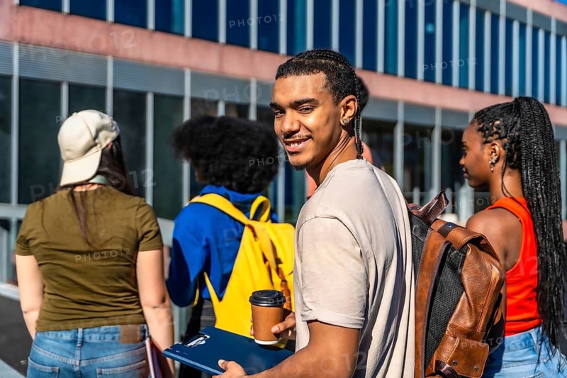 Diverse students walking together on campus, carrying backpacks and books while a male student smiles at the camera, capturing the essence of education, friendship, and student life