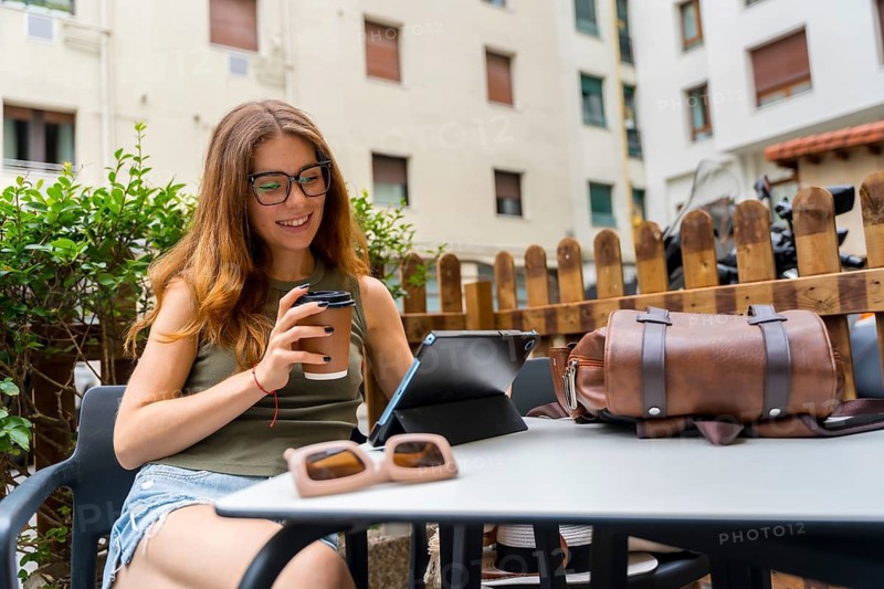 Young woman wearing glasses sitting at an outdoor cafe table, happily drinking coffee and using a digital tablet for remote work or studying in a city environment