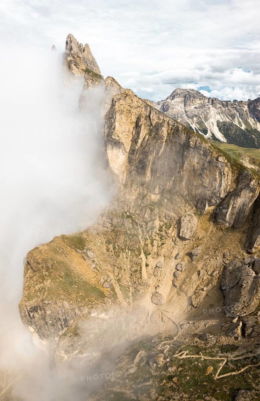 Dramatic drone aerial view jagged rock cliffs Seceda ridge line partly covered rolling white clouds switchback hiking trail valley alpine landscape South Tyrol Dolomites Italy