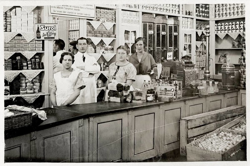 Colonial goods, sales counter, shop, business, profession, twenties, saleswoman, a group of people behind a counter surrounded by shelves full of canned goods, around 1928