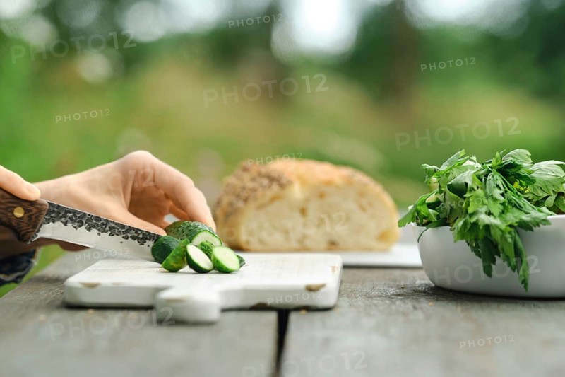 Person slices cucumbers on wooden cutting board. Fresh bread sits in background. Green herbs are nearby. Scene takes place outdoors in daylight with blurred greenery