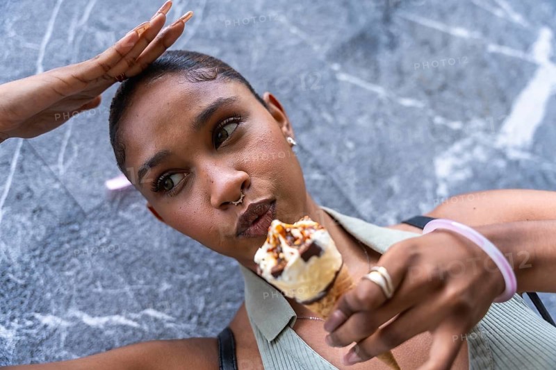 Young black woman holding an ice cream cone with anticipation, enjoying a sweet treat against a marble textured urban background, embodying casual summer style and city life pleasure