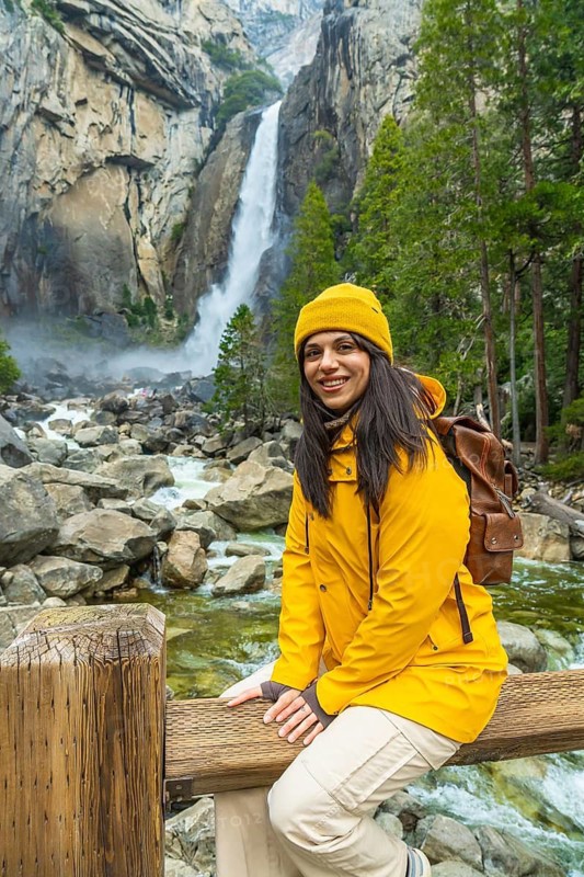 Woman smiling while sitting on a wooden fence, enjoying nature and experiencing the power of lower yosemite falls and the merced river crashing down granite cliffs in yosemite national park