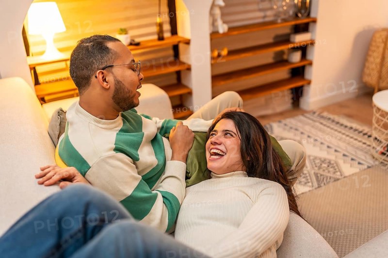 Young couple relaxed on a cozy sofa at home, sharing laughter and warm conversation in a bright living room, enjoying an intimate, candid moment of connection and joy