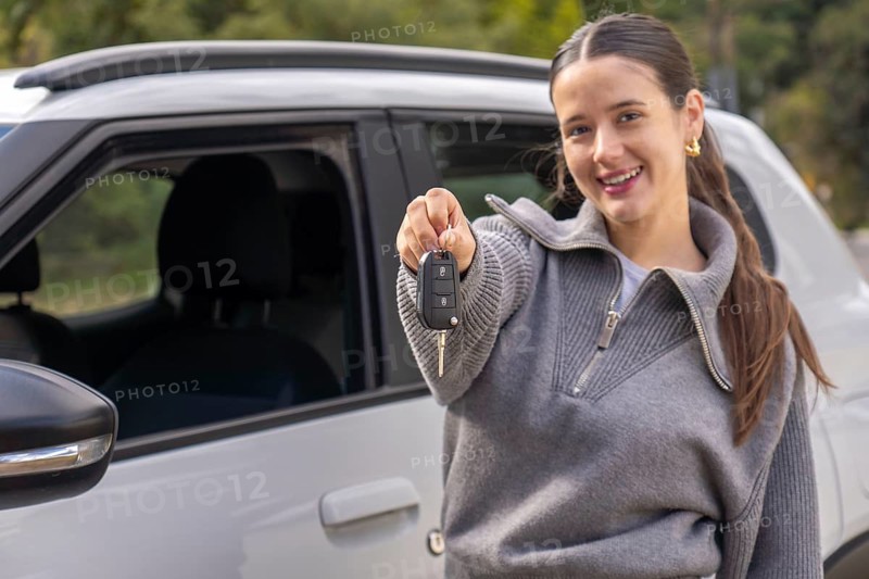 Close-up portrait of smiling young woman holding car key up to camera, blurred vehicle in background, ownership concept