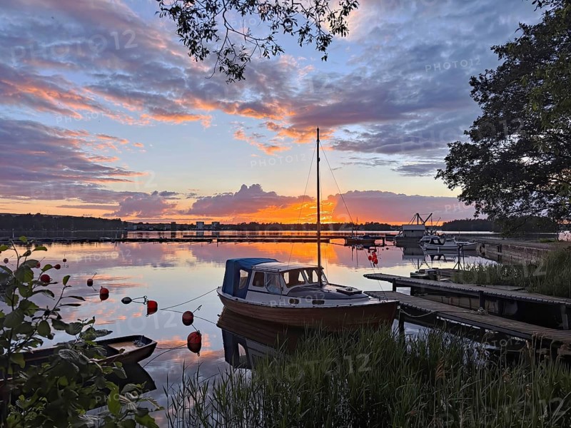 A boat is moored on a jetty in a calm lake at sunset with glowing clouds and reflections in the water, Göta Canal, Karlsborg, Västra Götalands län, Sweden