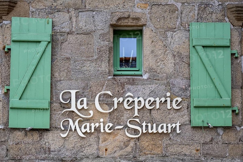 Close-up of an old Breton stone house made of grey granite with distinctive, open wooden shutters in light green, an ornate name of the Marie Stuart crêperie on the façade and a rustic building style in the historic city center. Roscoff, Finistère Department, Brittany, France