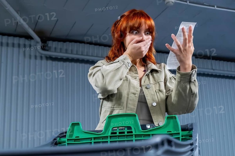 Young woman seeing her shocking grocery store bill, reacting with hand covering mouth, symbolizing rising inflation and increased cost of living impacting expenses