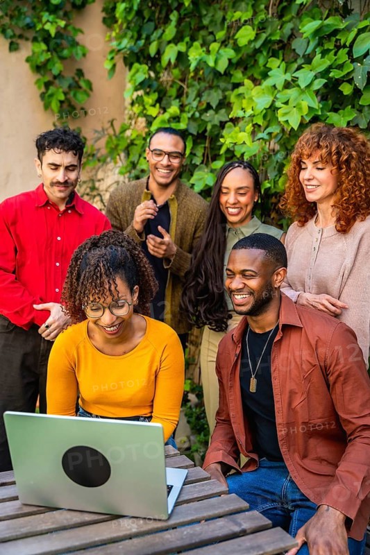 Group of diverse people collaborating and communicating during an outdoor therapy session, viewing information together on a laptop with smiles, embodying community support and teamwork