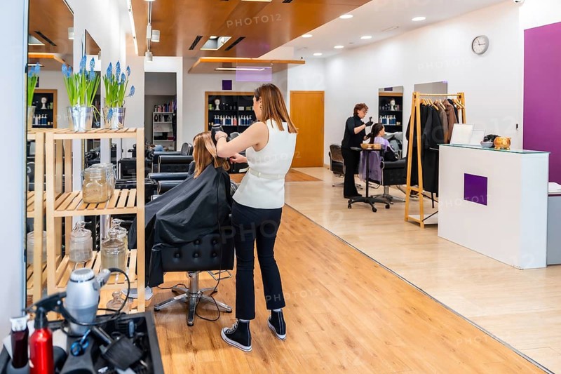Professional hairdresser working on a client's blow dry at a salon station, providing hair care services in a contemporary beauty studio environment with another stylist attending a customer