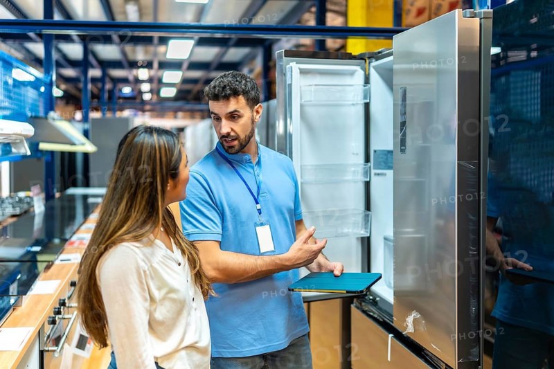 Sales assistant points at an open modern refrigerator while explaining features on a tablet to a female customer in a home improvement store, helping with her purchase decision