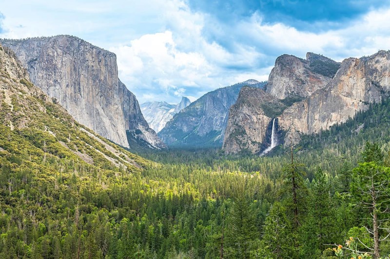Yosemite valley landscape showing el capitan cliffs, bridalveil fall cascading down granite walls, surrounding pine forest, and half dome under a dynamic blue sky with clouds