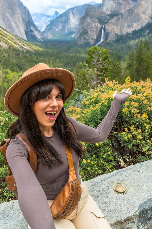 Woman smiling and gesturing at expansive yosemite valley from tunnel view, with bridalveil fall, el capitan and granite cliffs in a scenic travel and nature panorama