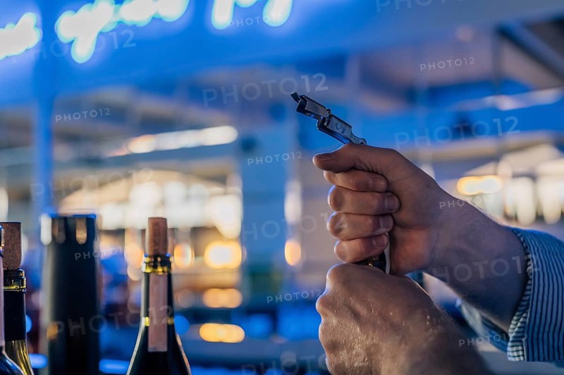 Men's hands open a bottle of wine at the bar. A close-up shows a sommelier's hands opening a wine bottle with a corkscrew. Several other bottles stand in the softly blurred blue background, creating a refined bar or restaurant atmosphere. The moment captures the precision and technique of wine service, highlighting preparation, hospitality, and evening mood