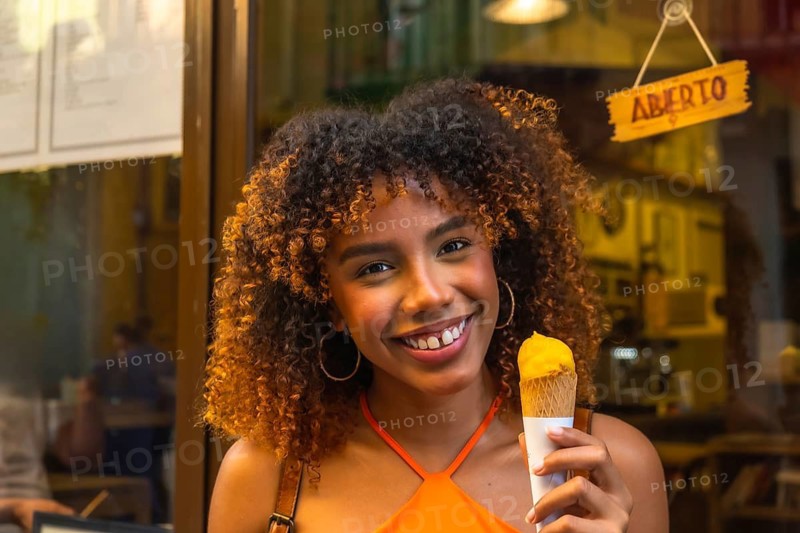 Young woman with curly hair smiling at camera while holding an orange gelato cone, enjoying a sweet, refreshing summer treat outside a storefront with an open sign