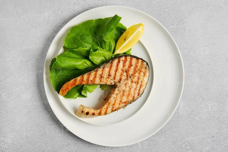Grilled salmon steak on white plate with salad and slice of lemon. This meal is prepared in kitchen and ready to eat. This dish shows simple cooking and fresh ingredients for lunch