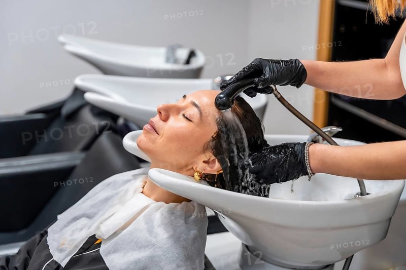 Female client relaxing with eyes closed while a professional hairdresser, wearing black gloves, washes her hair over a ceramic basin at a modern hair salon, focusing on hair care and wellness