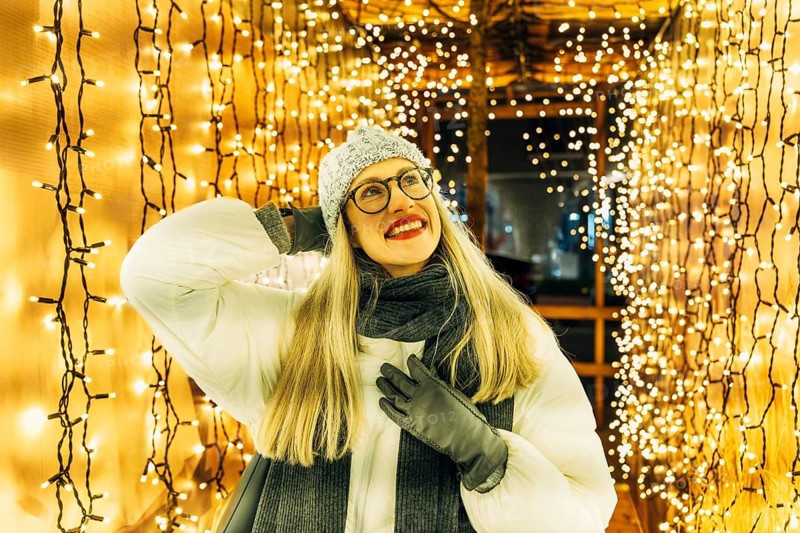 Young woman in glasses smiling in winter clothing under festive lights. A woman wearing a light winter coat, gray knit hat, scarf, gloves and glasses stands inside a tunnel of warm yellow string lights hanging vertically along the walls and ceiling. Her upward gaze and smile are illuminated by the dense festive glow, highlighting winter textures and the cozy holiday atmosphere of a nighttime seasonal display. Christmas atmosphere