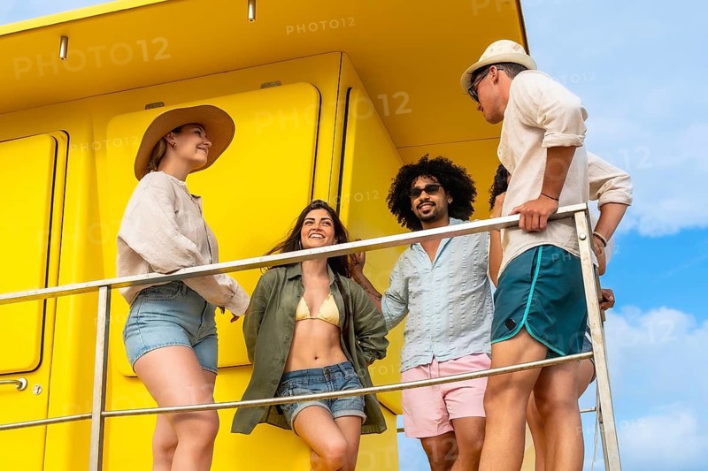 Group of diverse young adults laughing and talking, standing on a bright yellow lifeguard station enjoying their summer holidays under a clear blue sky