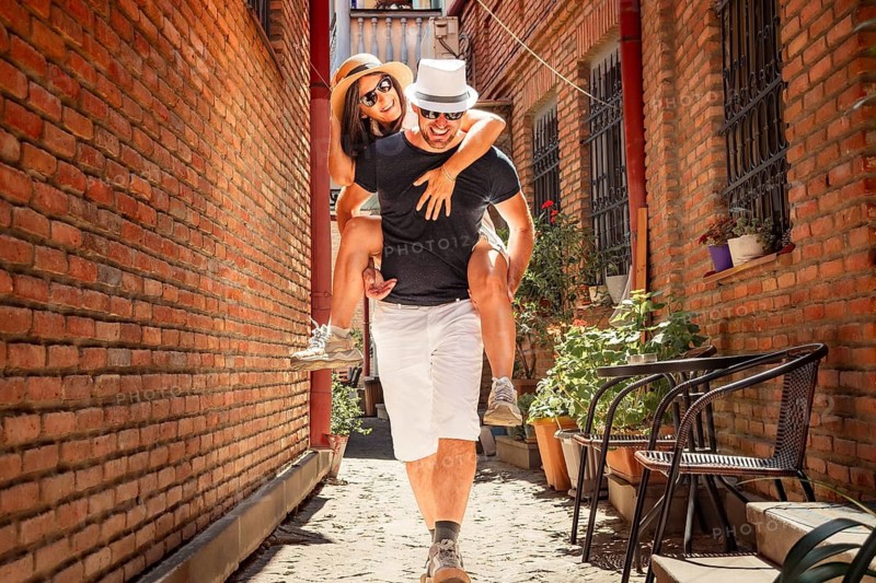 Beautiful young tourist couple Happy caucasian couple having fun piggyback visiting walk narrow alley street. Tourists enjoying holiday in Europe. Vacation and happy lifestyle concept