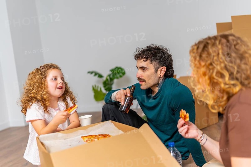 Family, including a father, mother, and daughter, sitting on the floor, enjoying pizza and drinks amidst moving boxes, celebrating their relocation into a new house