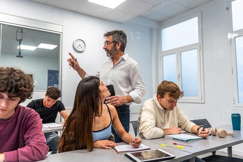 Teacher engaging female student at front of bright classroom while peers take notes and study attentively during a college lesson, focused, diverse group learning together