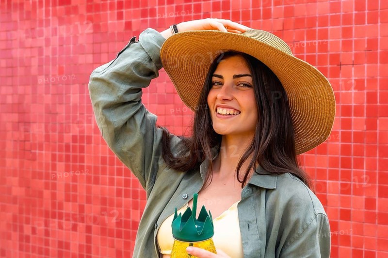 Young woman smiling while posing with a sun hat and a refreshing pineapple shaped drink, standing against a vibrant red tiled wall, embodying a cheerful summer lifestyle