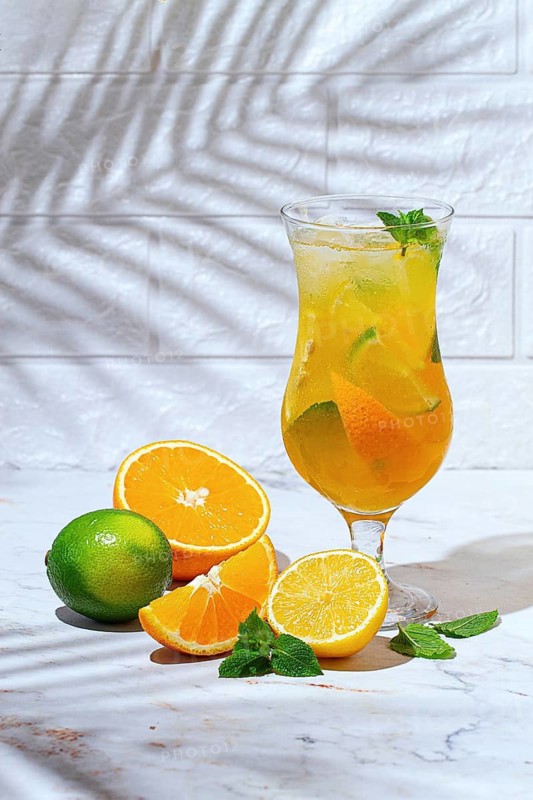 A glass with a drink made from orange and lime sits on a marble table. Fresh orange and lime slices are next to the glass, and there are mint leaves for garnish
