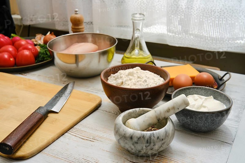 A kitchen scene shows fresh ingredients laid out on a wooden table. There are tomatoes, flour, eggs, and cooking tools. Olive oil and spices are also visible, suggesting meal preparation