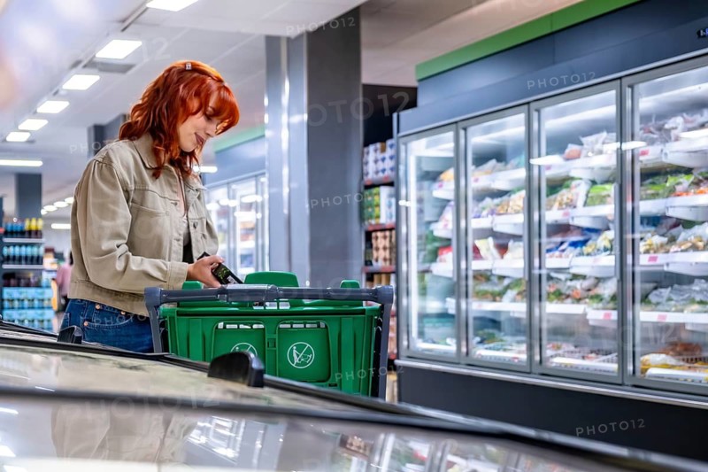 Red haired woman pushing a green shopping cart while selecting frozen and fresh items from a refrigerated display in a modern supermarket aisle, everyday grocery shopping scene