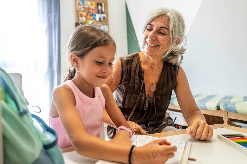 Grandmother sitting at a table, smiling and supervising her granddaughter coloring in a book, fostering learning, bonding, and family connection in a casual home environment