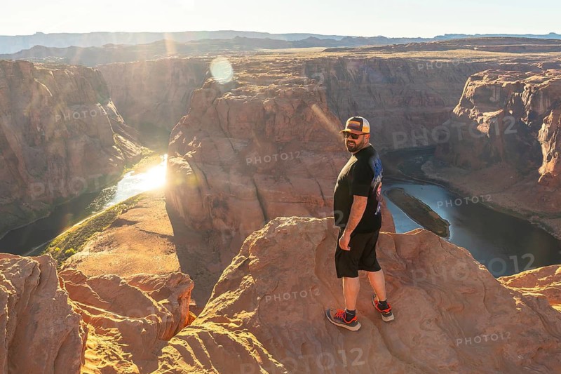 Man stands on a cliff edge overlooking horseshoe bend and the colorado river at golden hour, warm light on sandstone meanders and expansive arizona canyon landscape